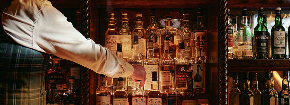 Warmly lit Bertie's Bar in The Fife Arms, Braemar Scotland, filled with stacked rows of Scotch whisky bottles, showing a bartender in a tartan vest reaching for a bottle on a shelf.
