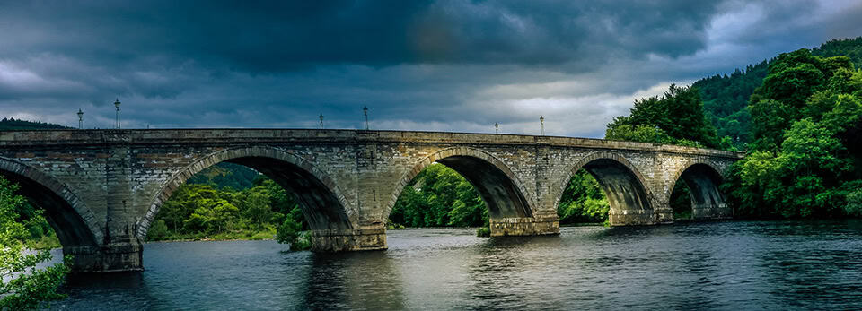 Stone arched bridge in Dunkeld, Scotland spanning a the river Tay under a dramatic, dark cloudy sky, flanked by dense green trees on both sides.