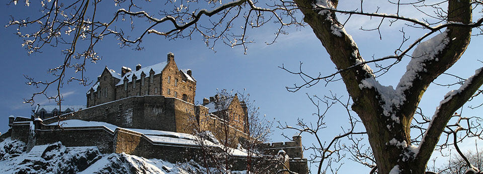 Low-angle shot of snow-covered Edinburgh Castle on a rocky hill under a clear blue sky, framed by bare, snowy tree branches.