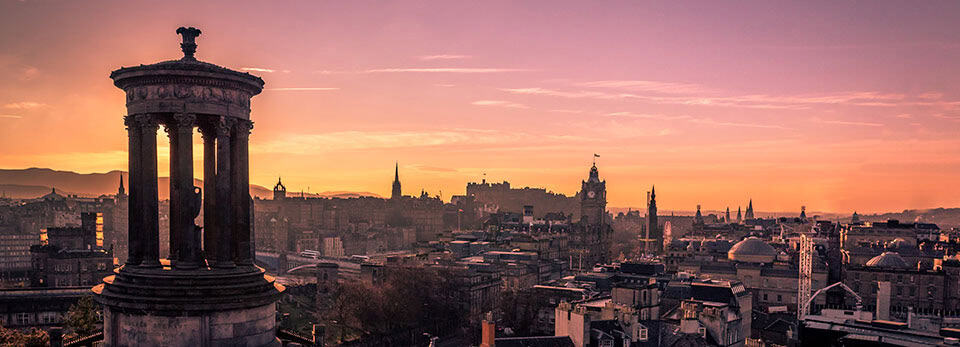 Panoramic sunset view over Edinburgh's skyline, featuring the Dugald Stewart Monument in the foreground against a warm, orange-pink sky.