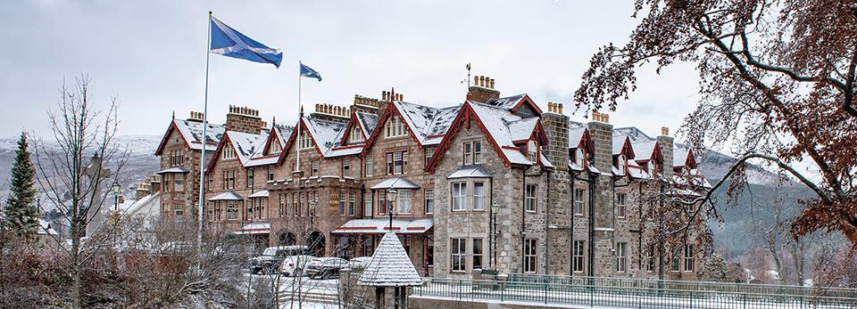 Large, historic stone The Fife Arms Hotel decorated with red trim and Scottish flags, surrounded by snow and bare trees with snow-capped hills in the background.