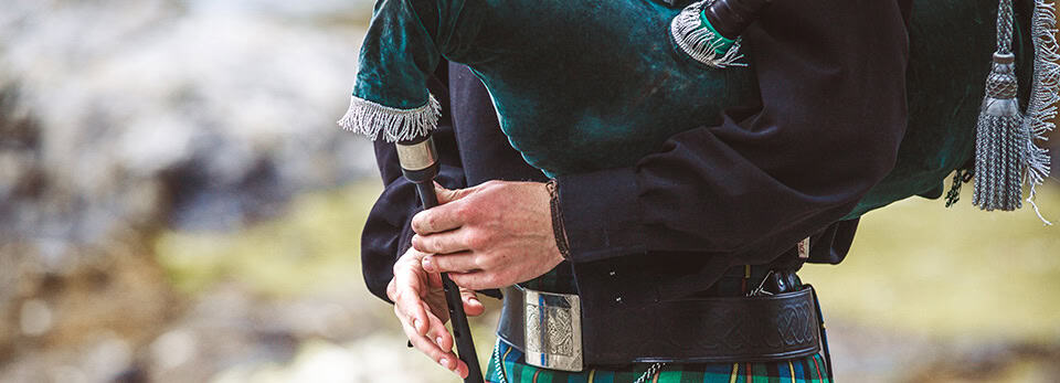 Close-up of a person wearing a traditional kilt and black jacket playing the bagpipes, holding the wooden chanter.