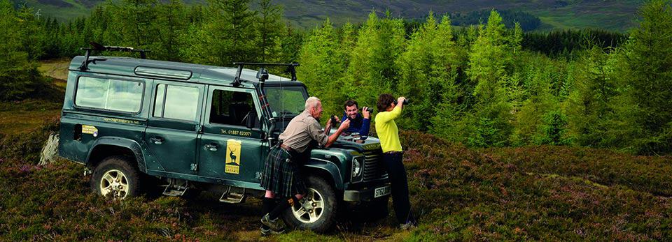 A green Land Rover Defender parked in a lush, wooded heath, with three people—one wearing a kilt—leaning against the front and using binoculars.