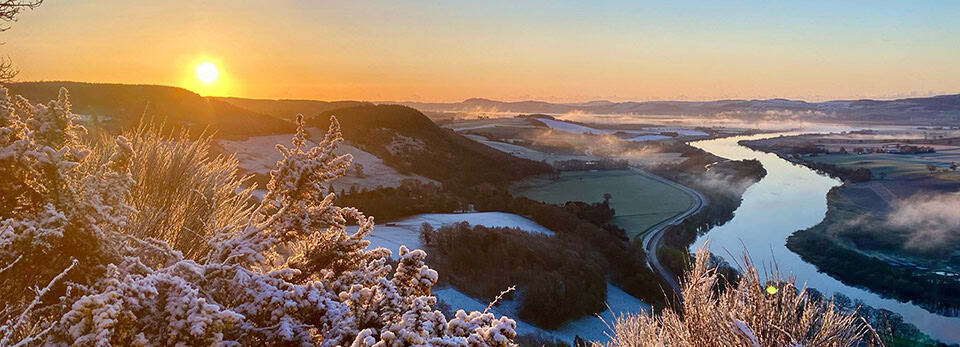 Panoramic view at sunrise over a frosted valley, featuring frost-dusted gorse bushes in the foreground, a winding river, and rolling snow-covered hills under a warm, glowing sky.