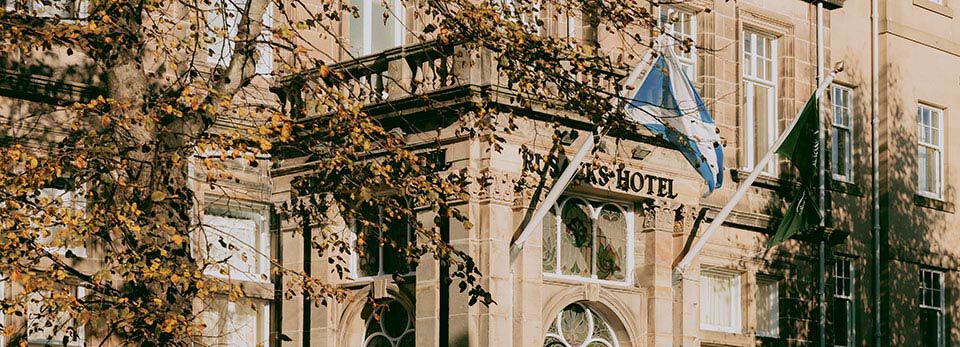 Exterior of the sandstone Rusacks Hotel in St Andrew's building framed by autumn trees with yellow leaves, featuring a Scottish Saltire flag above the main entrance.