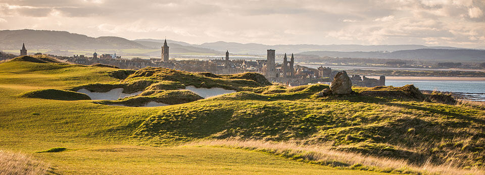 Scenic view across a St Andrew's golf course with grassy dunes towards a St Andrews, a Scottish coastal town, historic ruins, and snow-capped mountains.