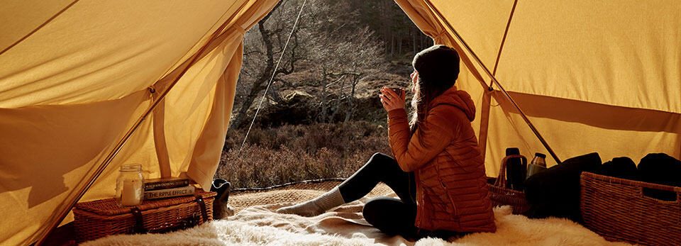 Cosy view from inside a brightly lit bell tent looking out onto a Braemar heathland landscape in Scotland, as a person wrapped in a jacket holds a warm mug while seated on blankets.